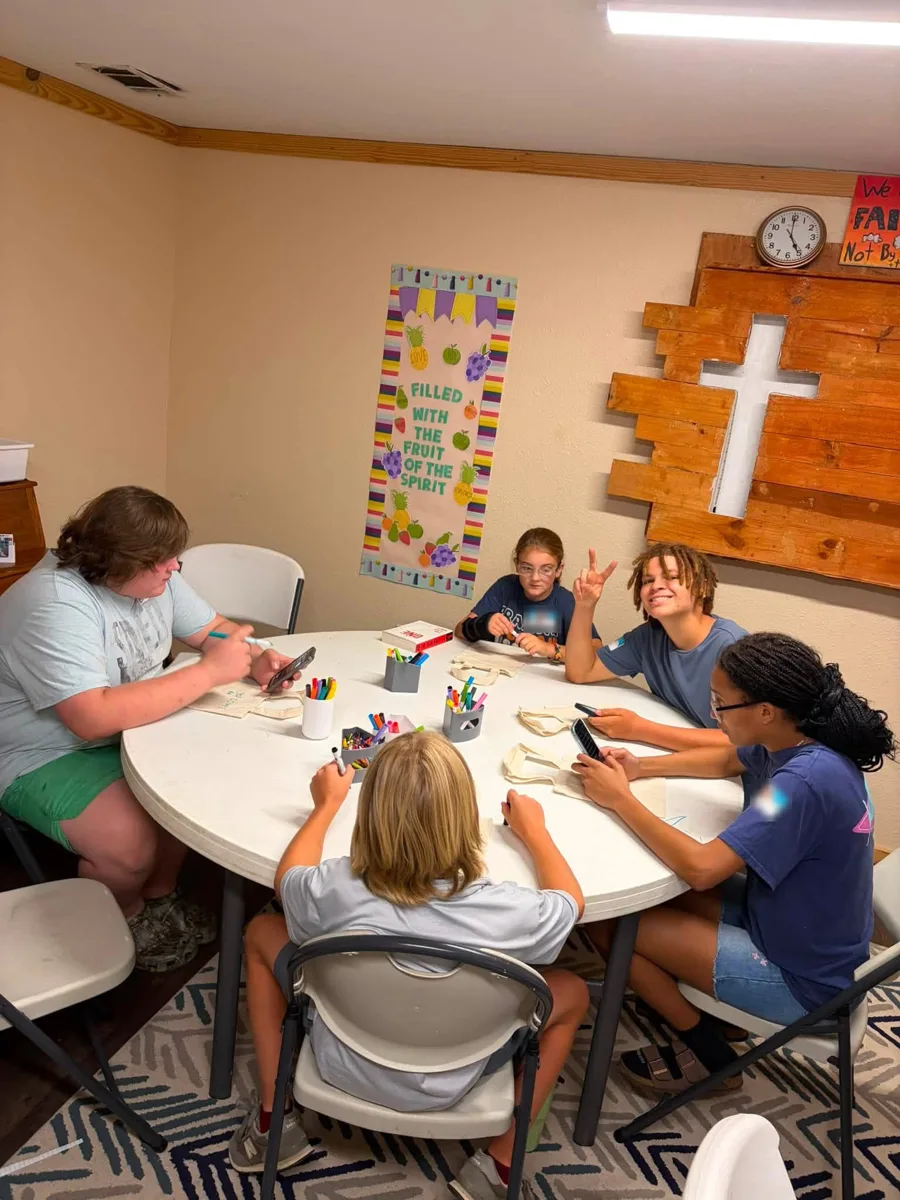 Students gathered around a table with markers and a Bible, beneath a wooden cross and a Fruit of the Spirit poster