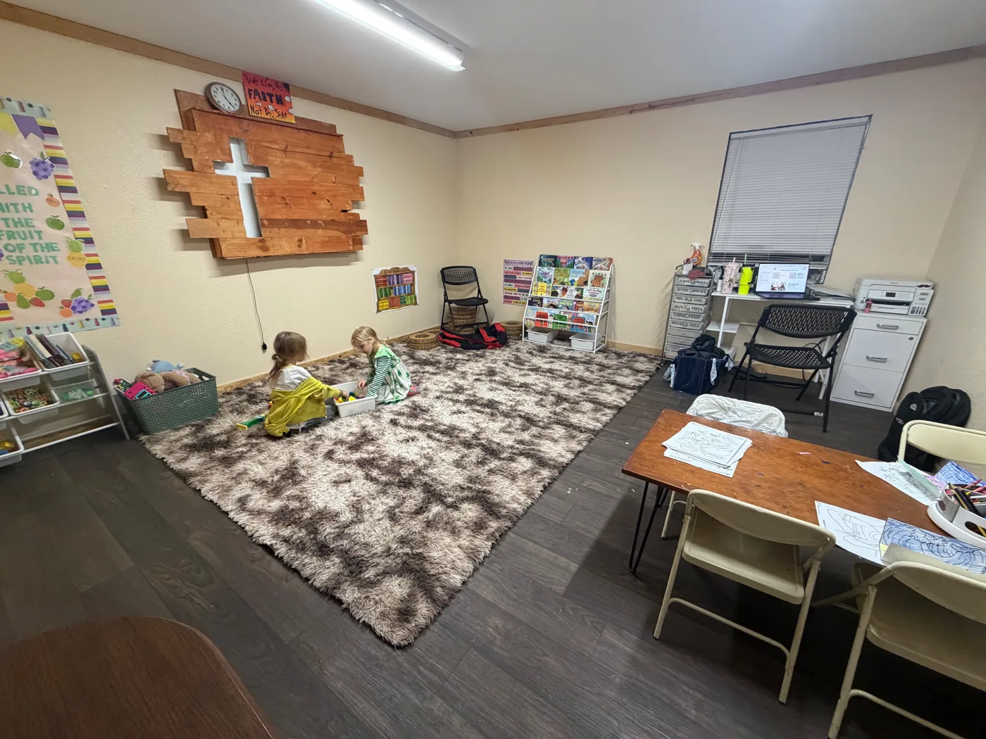 Two children playing on a cozy rug in the Children’s Church room, with a wooden cross and Bible bookshelf on the wall