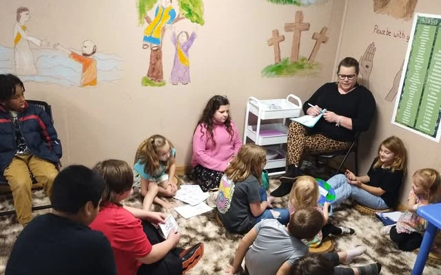 Children gathered on the floor around their teacher for a Wednesday night lesson, with Bible story paintings on the walls